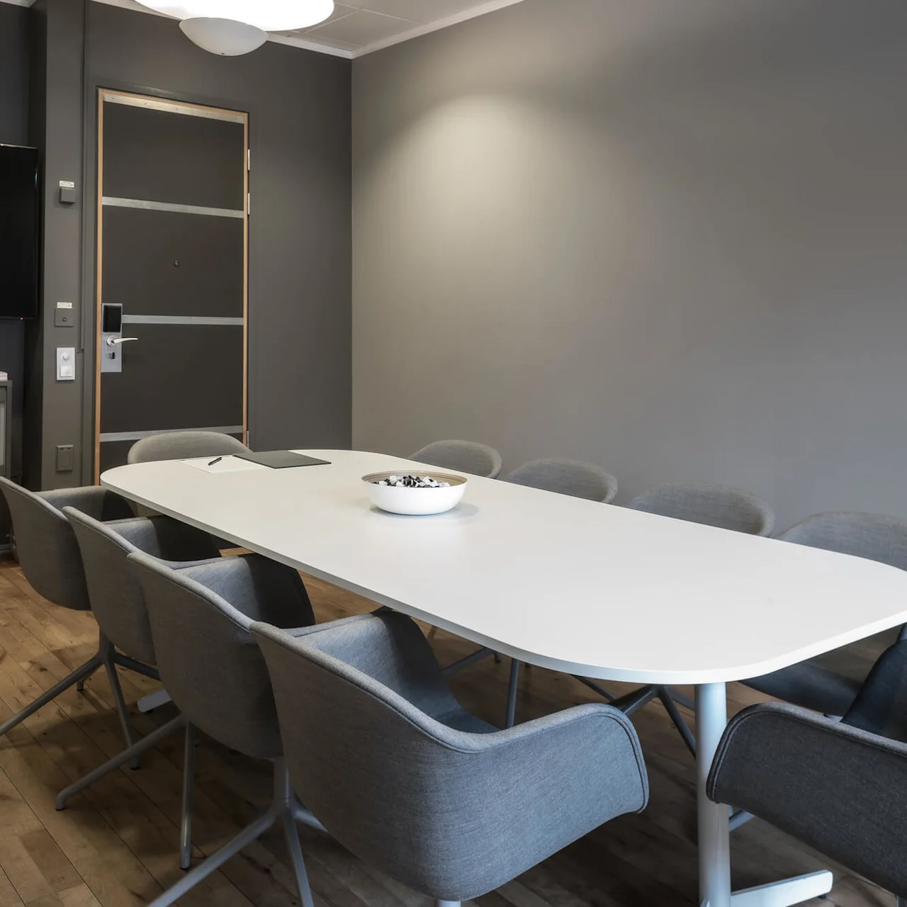 A long white table with eight gray chairs surrounds it in a modern conference room. A flat-screen TV hangs on the gray wall beside a cabinet, under soft lighting.