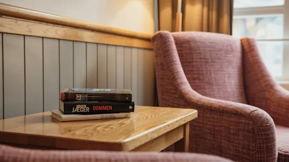 A cozy reading nook with two armchairs, a side table with books, including one titled JAEGER DOMMEN, and a lamp.