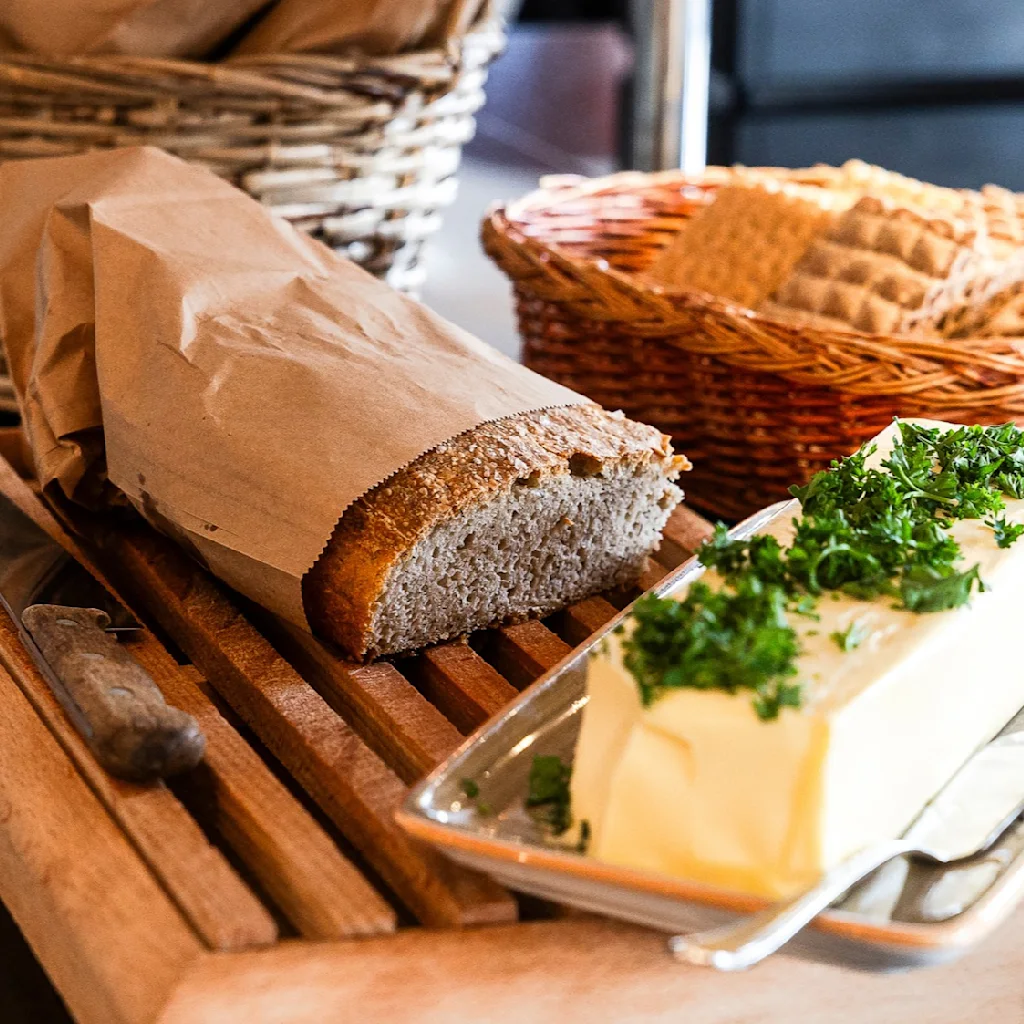 Brød i papirpose ligger på et skjærebrett ved siden av en smørfat med urter. I bakgrunnen står en kurv med kjeks.
