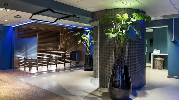 A spa area features large potted plants standing beside a glass-walled sauna. Warms lights illuminate the wooden interior, and towels are neatly stacked in the background.