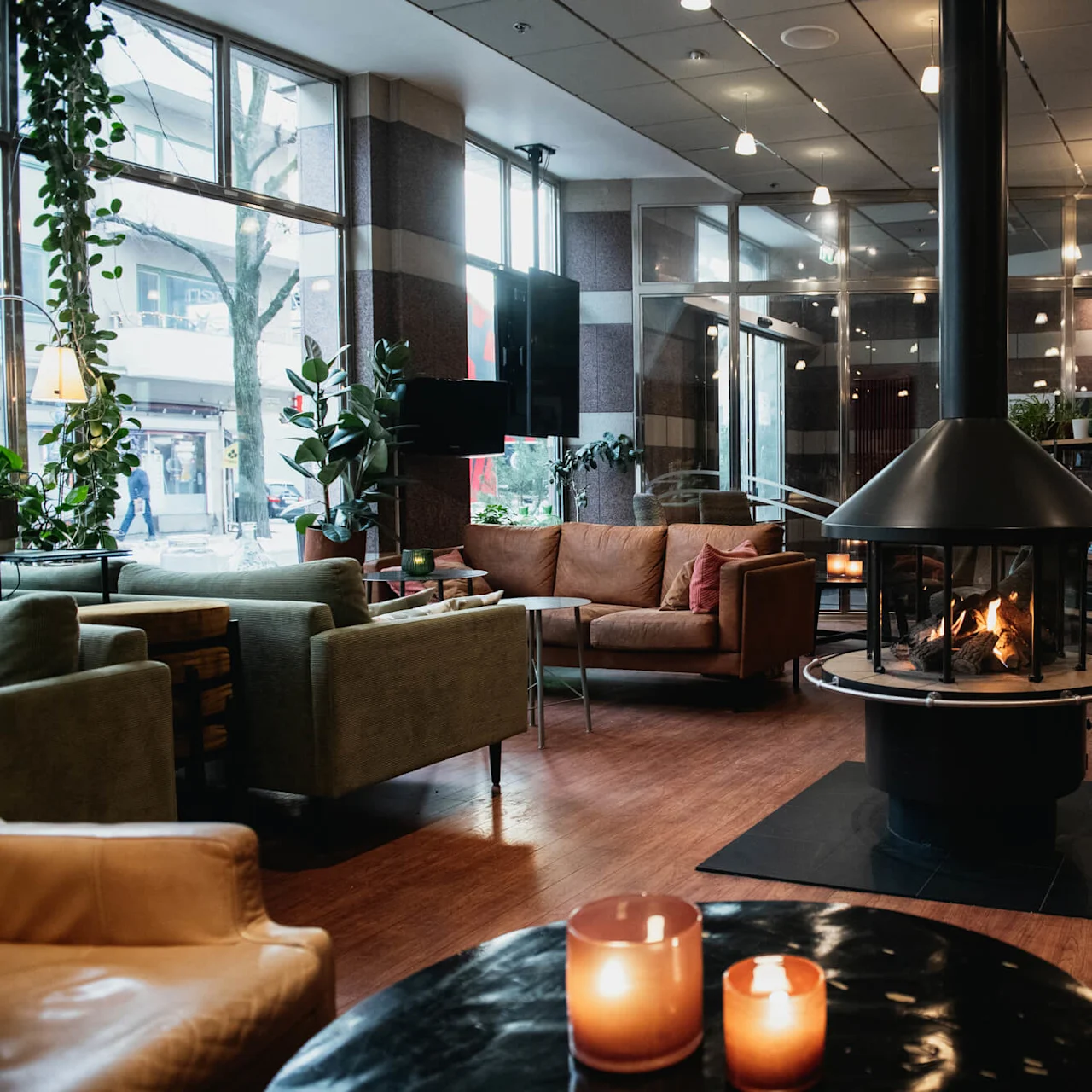 A cozy lounge with a central fireplace surrounded by sofas and chairs. Large windows provide natural light, and candlelit tables add warmth. Potted plants decorate the modern setting.