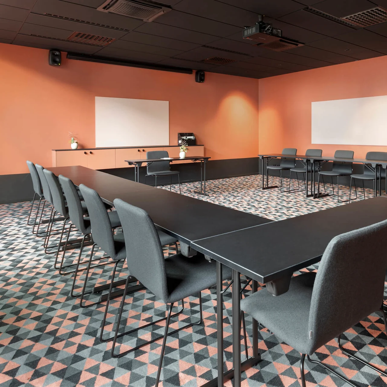 Modernt konferensrum med U-formade svarta bord och grå stolar, på en geometriskt mönstrad matta. Orange väggar med två stora whiteboards och en takmonterad projektor.