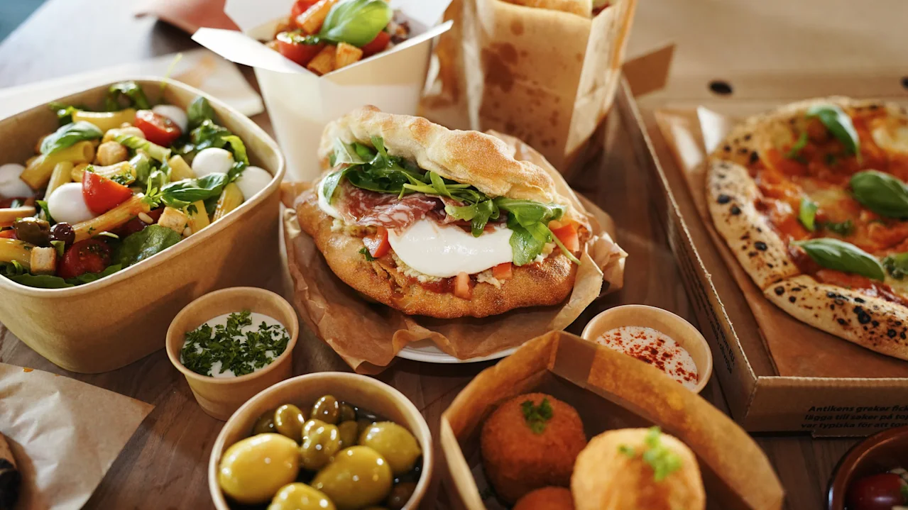 Erilaisia italialaisia ruokia, kuten pizzaa, salaattia, voileipiä ja oliiveja, on asetettu puupöydälle, mikä viittaa rentoon ruokailu- tai takeout-tilanteeseen.