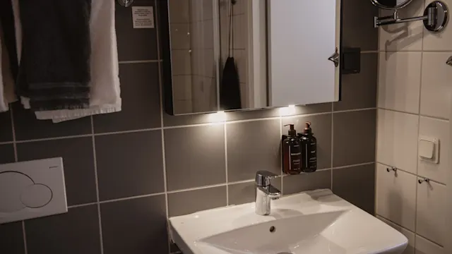 En vit tvättställ under en upplyst spegel, monterad på en kaklad badrumsvägg, med handdukar, behållare och en spolknapp i närheten. Ingen text synlig eller närvarande.