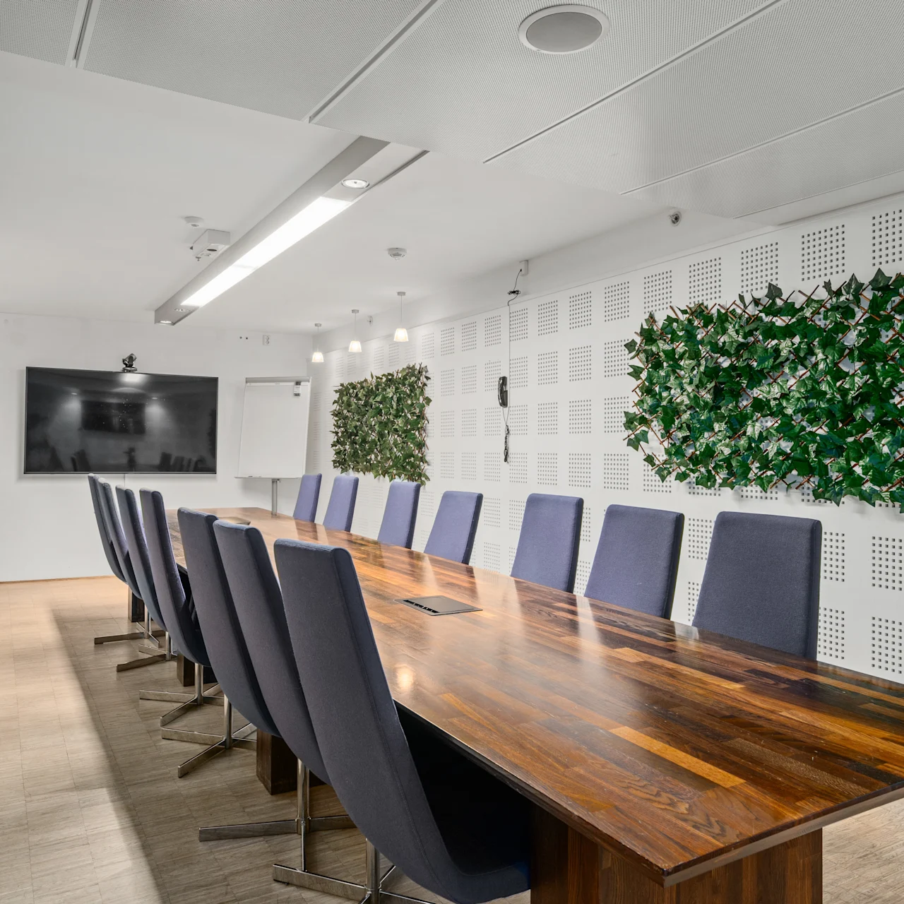 Ett långt konferensbord av trä omges av stolar i ett modernt mötesrum med en väggmonterad TV och dekorativa växter på väggarna.