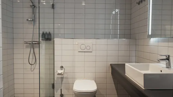 Valoisa, moderni kylpyhuone, jossa on valkoiset kaakeloidut seinät, lasitettu suihku, wc ja allaskaappi peilillä.