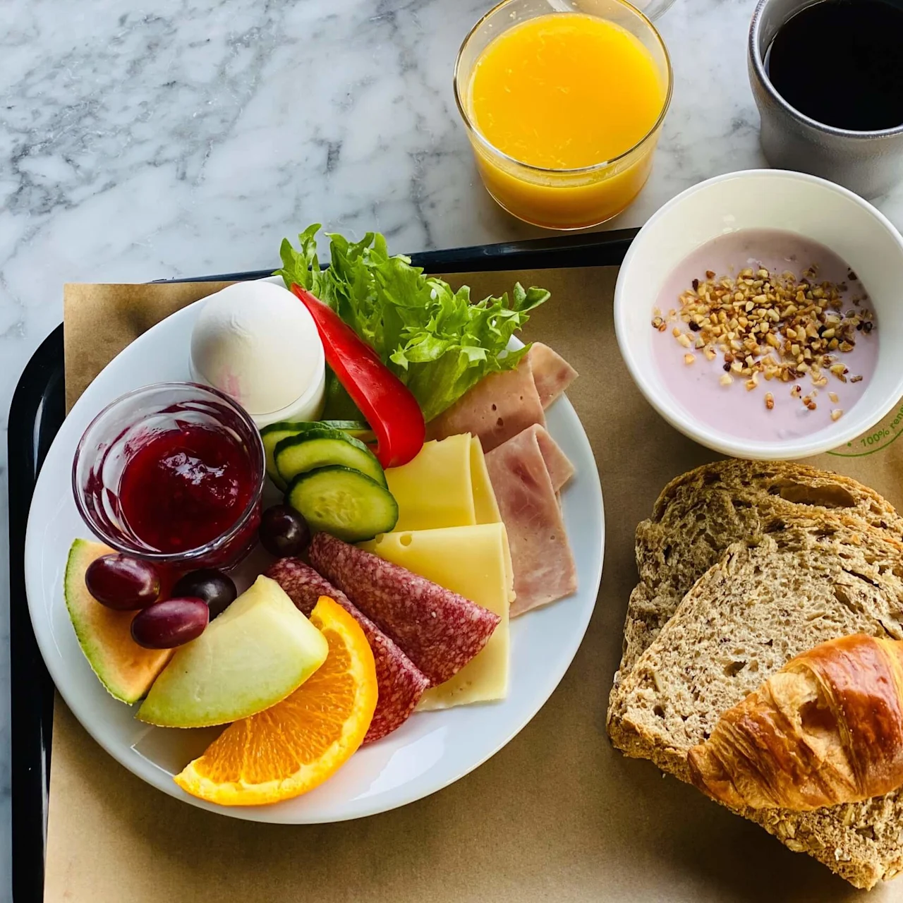 En tallerken med ost, skinke, salami, frugt og grøntsager står på en bakke ved siden af yoghurt, en croissant, brødskiver, appelsinjuice og kaffe. Placeret på et marmorbord.