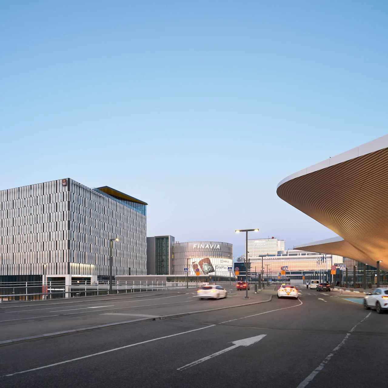 A modern building with a patterned facade stands by a road; cars pass by under a clear blue sky. The building is labeled "FINAVIA."