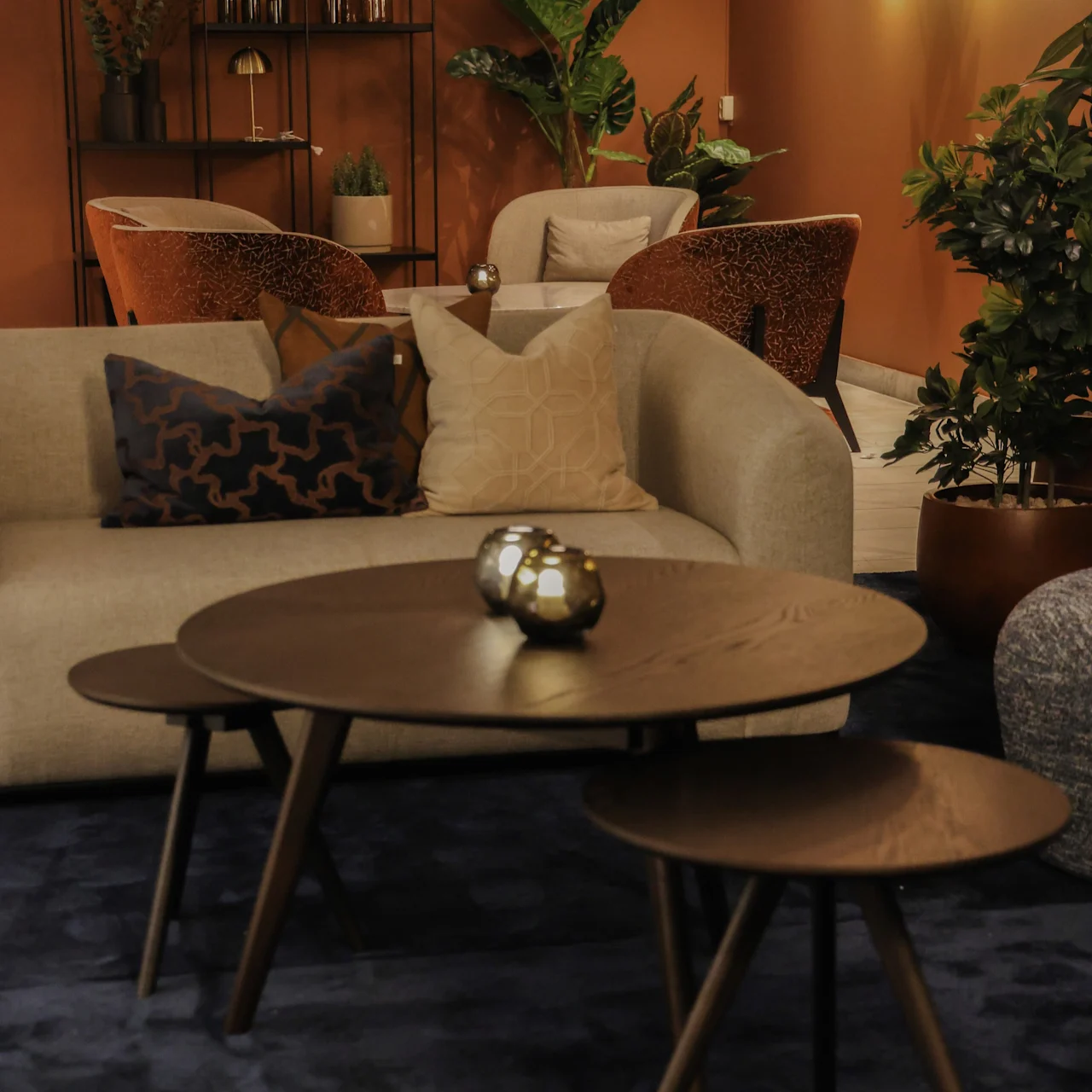 A round wooden coffee table set rests on a blue rug in a cozy living room. Nearby, a cream sofa with cushions and plants create a warm atmosphere.