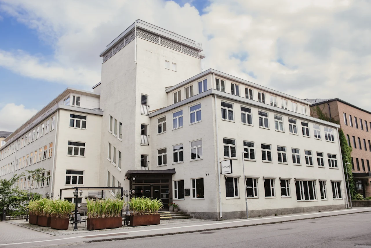 Exterior of a white hotel building with 'home hotel' sign, green plants, and a street in front. Home Hotel Uppsala.