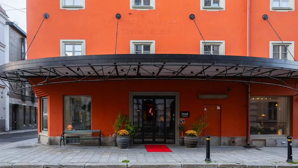 En levende orange bygning med store vinduer har en overdækket indgang med en rød løber. Potteplanter flankerer døren, og skilte siger "hotel." Det ligger på en brostensbelagt gade.