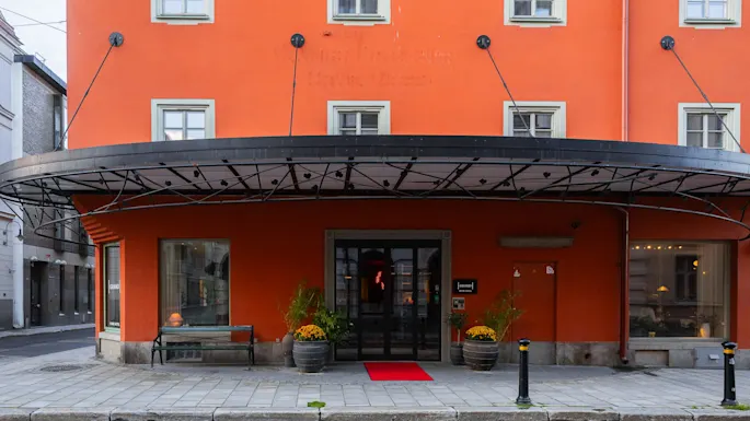 En lysende oransje bygning med store vinduer har en overbygd inngang med en rød løper. Potteplanter flankerer døren, og skilt sier "hotell." Det ligger på en brosteinsgate.