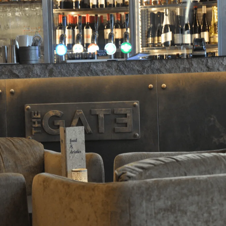 Mukavat tuolit baarin edessä, seinällä kyltti jossa lukee "THE GATE". Konteksti on sisätilan lounge-alue tai pubi.