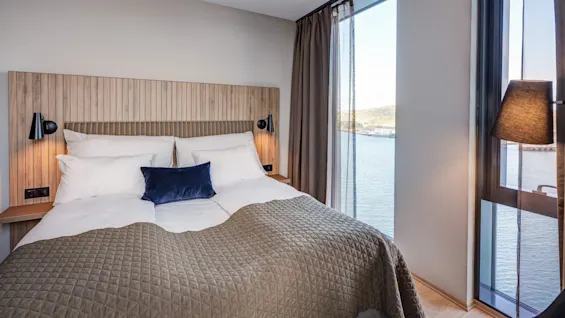 A bed with a brown quilted blanket and white pillows is positioned next to a large window overlooking a body of water, and is flanked by two black lamps.