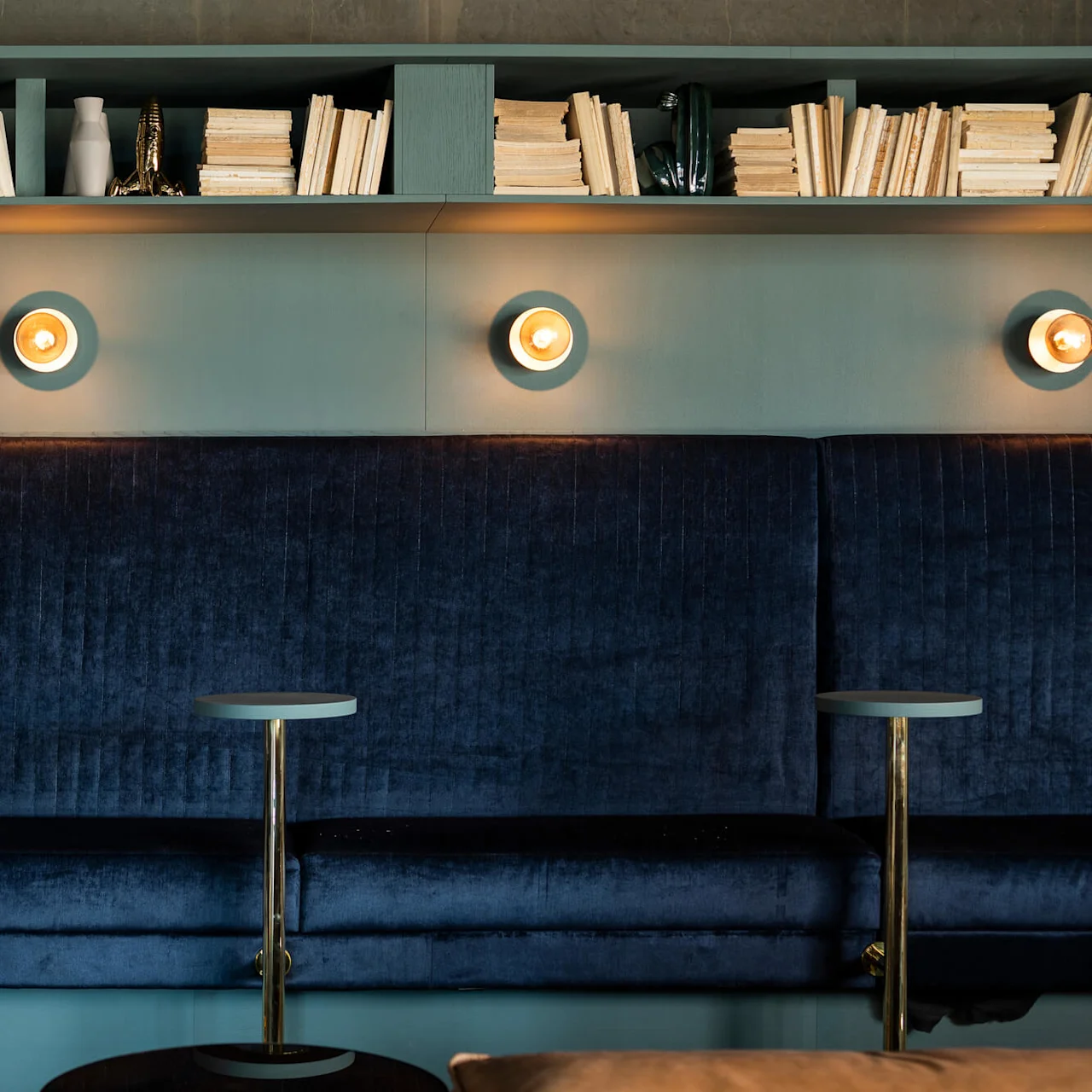 A deep blue velvet bench is situated against a wall with three round lights above. Shelves filled with stacked books and decorative items are above the lights.
