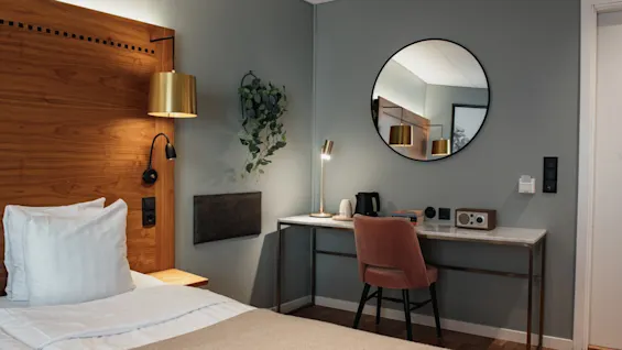 Et moderne hotelværelse med en seng, en sengegavl i træ og et skrivebordsområde med stol, spejl og varm messingbelysning.