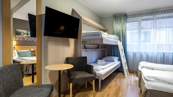 Bunk bed with folded towels is against a wall in a hotel room. Two chairs and a round table face a mounted TV. Window with curtains filters light.