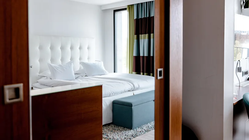 A neatly made bed with white linens and a tufted headboard stands in a bright room. Large window with dark curtains and a small bench at the foot.