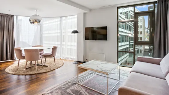 A modern living room with a dining area, hardwood floor, and sliding balcony door. Bright natural light illuminates the space.