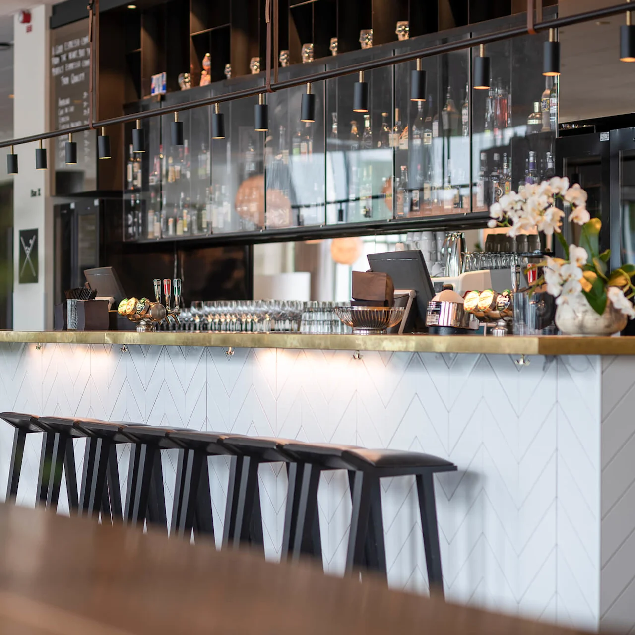Bar counter with rows of black stools. Glassware and flowers sit on the counter, with bottles and muted lighting overhead. Open, modern restaurant setting in the background.
