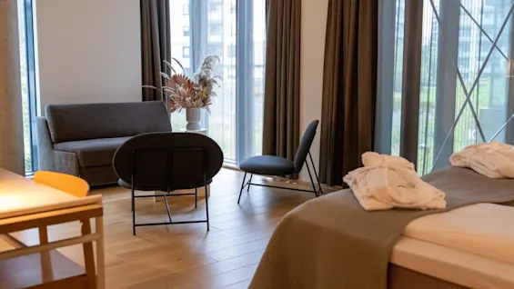 Moderne hotellrom med en koselig sittegruppe, store vinduer og en seng med brettede badekåper.