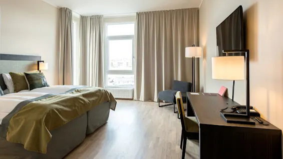 Siististi järjestetty hotellihuone, pedattu sänky, työpöytä tuoleineen, läppäri ja puhelin, sekä ikkuna kaupunkinäkymällä.