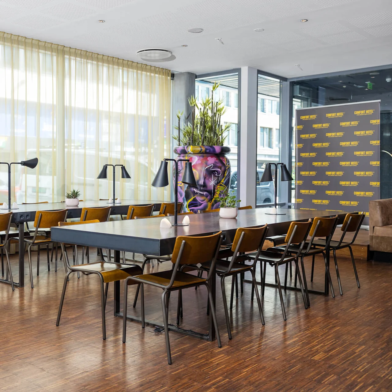 Tables lined with lamps and chairs in a brightly lit room featuring large windows and plants. A backdrop displays "COMFORT HOTEL™" repeatedly.