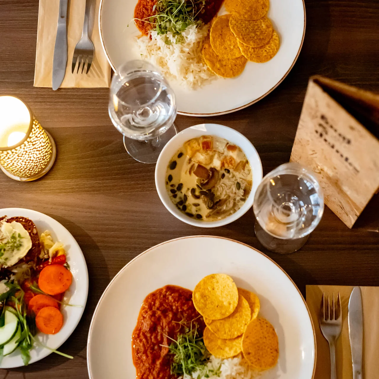 Ruokalautasia tarjoillaan tummalla puisella pöydällä yhdessä aterimien, lautasliinojen, vesilasien ja sytytetyn kynttilän kanssa.