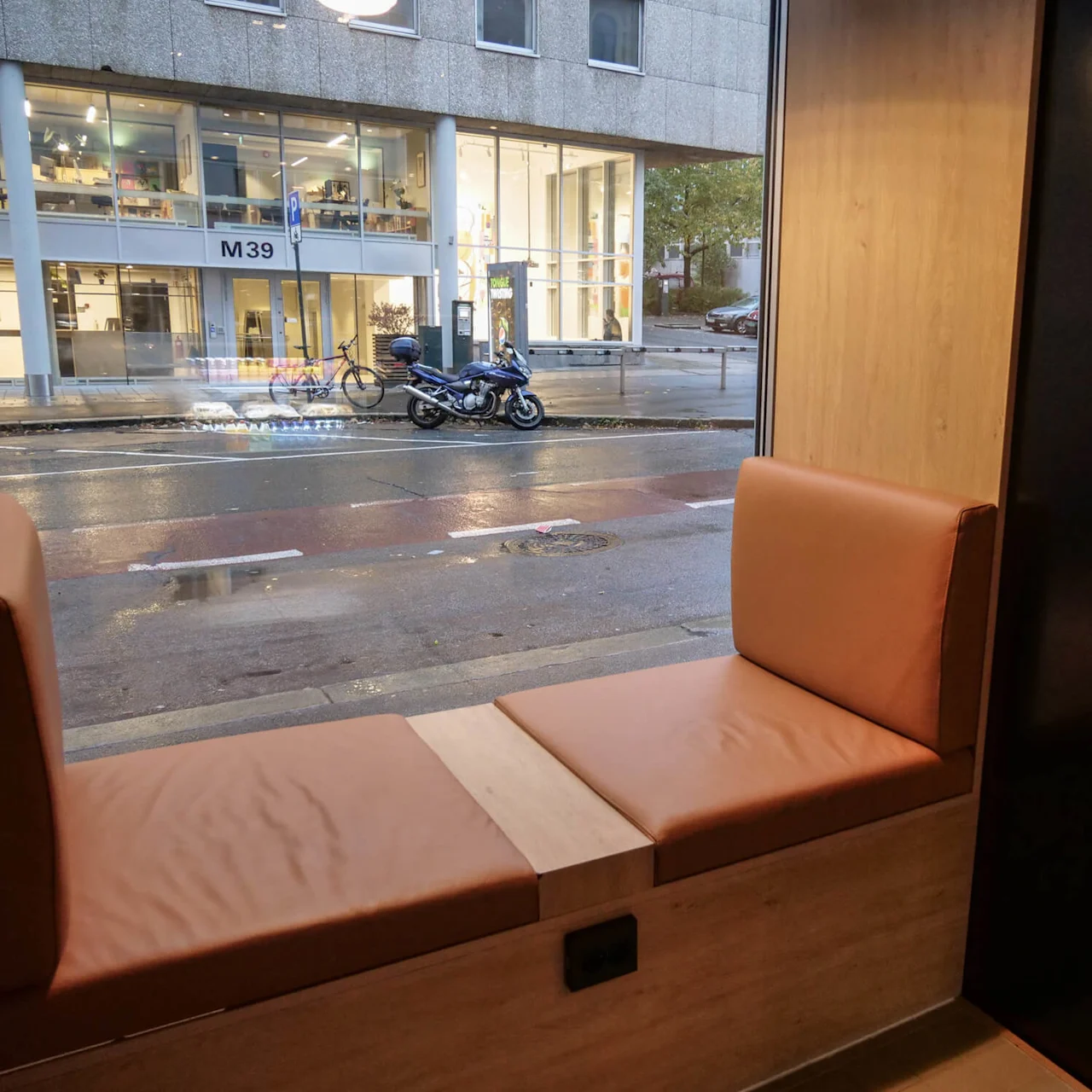 Cozy brown bench seating by a large window overlooking a city street with a parked motorcycle; across the street is a modern building labeled "M39" with glass windows.