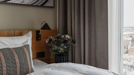 Et moderne hotellrom med en komfortabel seng, blomster ved sengen og et stort vindu med utsikt over byen.