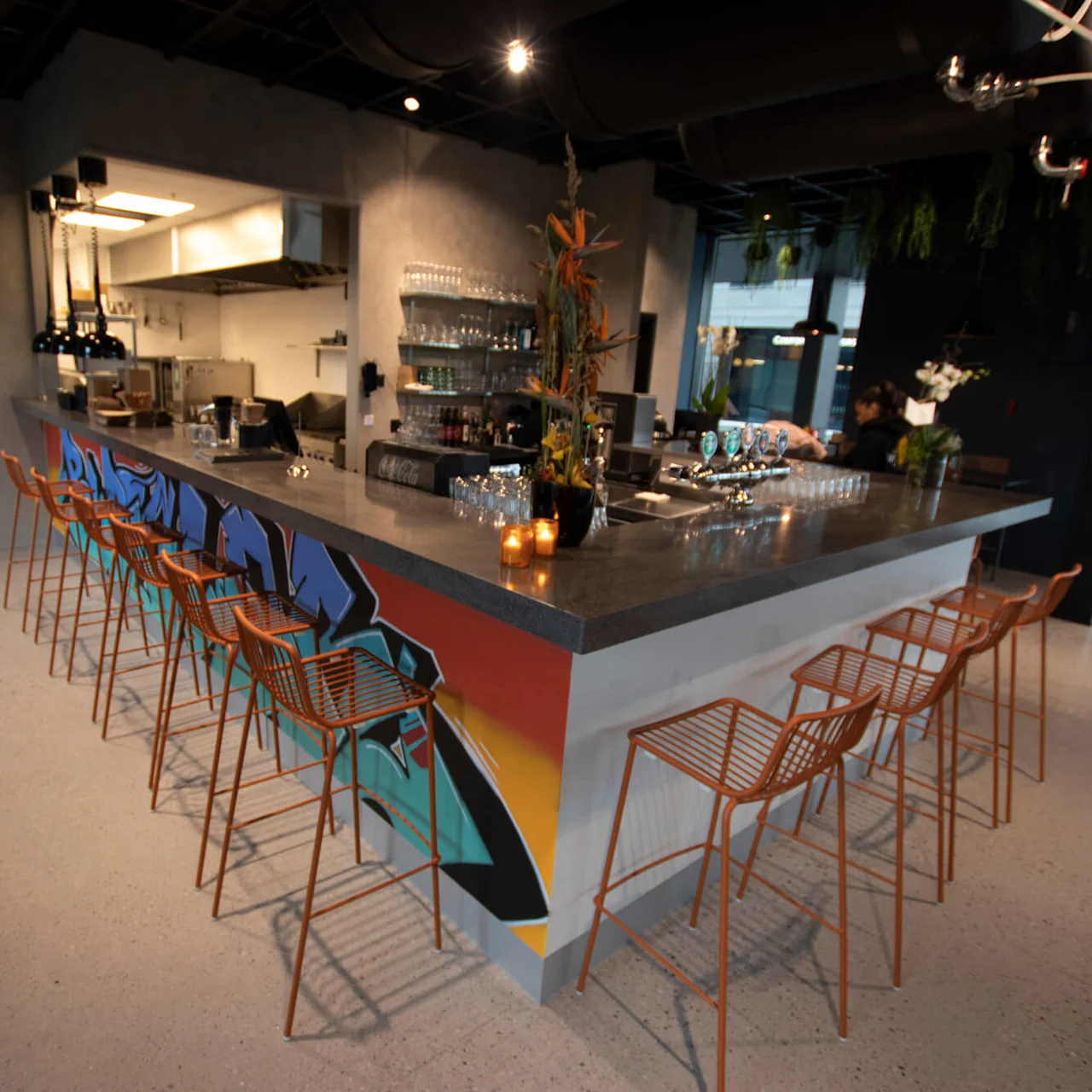 En bardisk med dekorativ graffiti har orange stole og er omgivet af moderne indretning, planter og køkkenudstyr i en café i industriel stil.