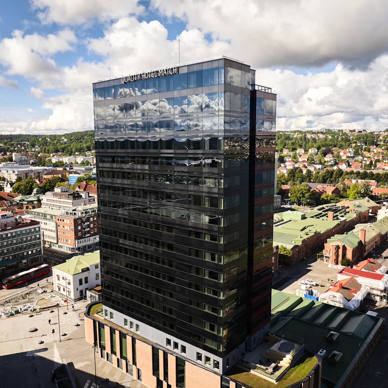 En høj, moderne glasbygning mærket "QUALITY HOTEL MATCH" reflekterer himlen, omgivet af bymiljø med træer og huse i det fjerne.