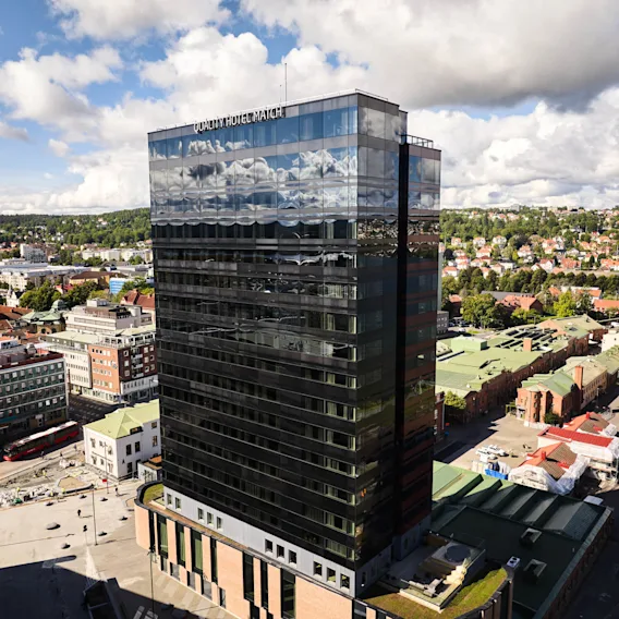 En høy, moderne glassbygning med påskriften "QUALITY HOTEL MATCH" reflekterer himmelen, omgitt av bylandskap med trær og hus i det fjerne.