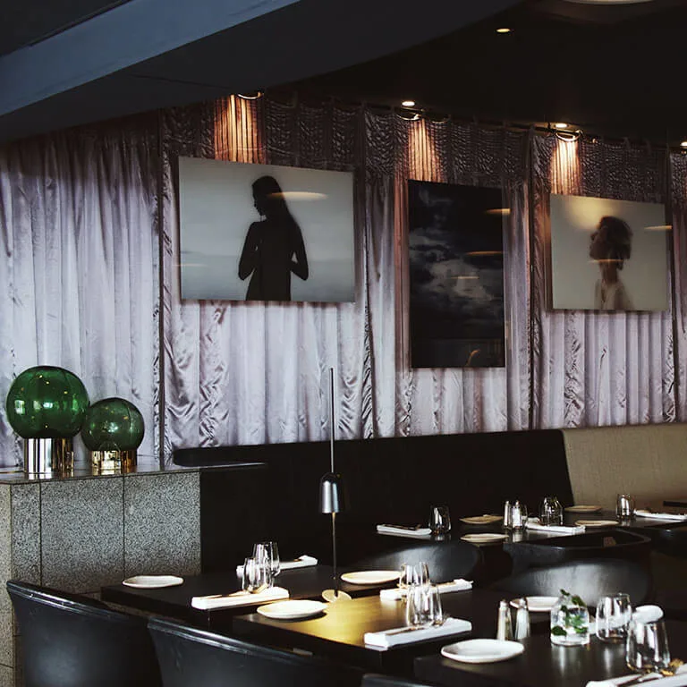 Spisebordene er pænt dækket med tallerkener og glas, mod en baggrund af indrammede silhuetfotografier på et draperet gardin i en svagt oplyst, moderne restaurant.