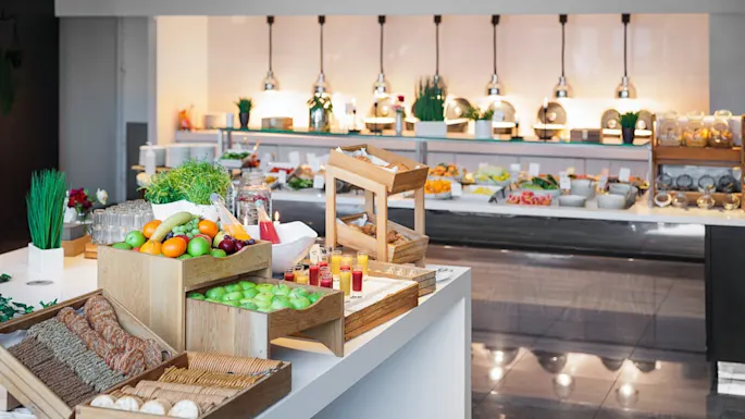 A buffet table displays fresh fruits, pastries, and juices on wooden trays in a well-lit dining area with modern decor and additional food options in the background.