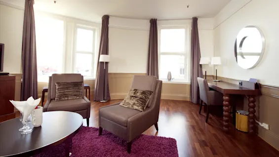 Et godt oplyst, rummeligt værelse med to lænestole, et sofabord og et skrivebord, med store vinduer og elegant, neutralt tonedekoration.