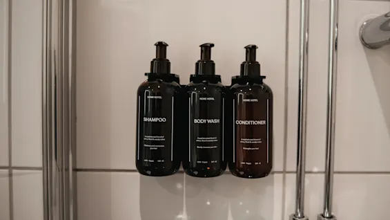 Tre mörka flaskor märkta SHAMPOO, BODY WASH och CONDITIONER är monterade på en vitkaklad duschvägg.