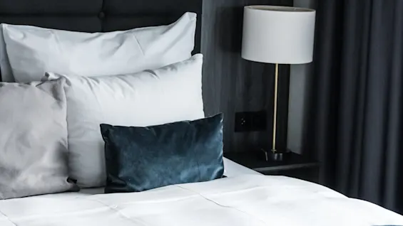 En seng med pent arrangerte puter, inkludert en mørkeblå, er i et moderne soverom med dempede toner. En lampe står på et nattbord ved trukne gardiner.