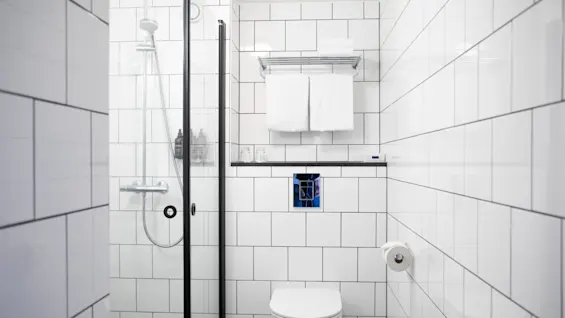 Et hvidt flisebelagt badeværelse med toilet ved siden af en glasindkapslet bruser. To håndklæder er pænt stablet på en kromhylde over toilettet, med en toiletpapirholder på højre væg.