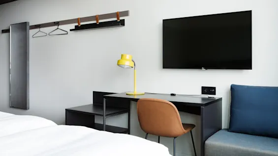 Ett skrivbord med en gul lampa står mot en väggmonterad TV. I närheten finns en stol och en spegel i ett minimalistiskt rum med en vit säng.