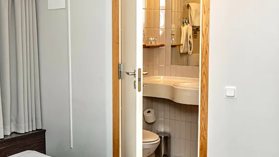 Ett rent och funktionellt hotellrum med en säng i förgrunden och en öppen dörr som visar ett väl upplyst badrum.