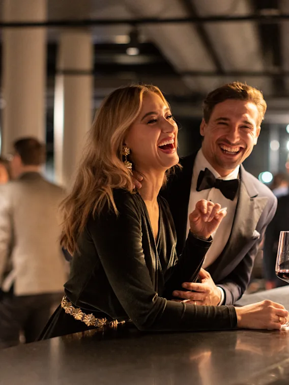 En kvinne og en mann, elegant kledd og leende, står ved en bar med drinker; bakgrunnen viser en livlig sosial sammenkomst med folk som mingler under dempet belysning.