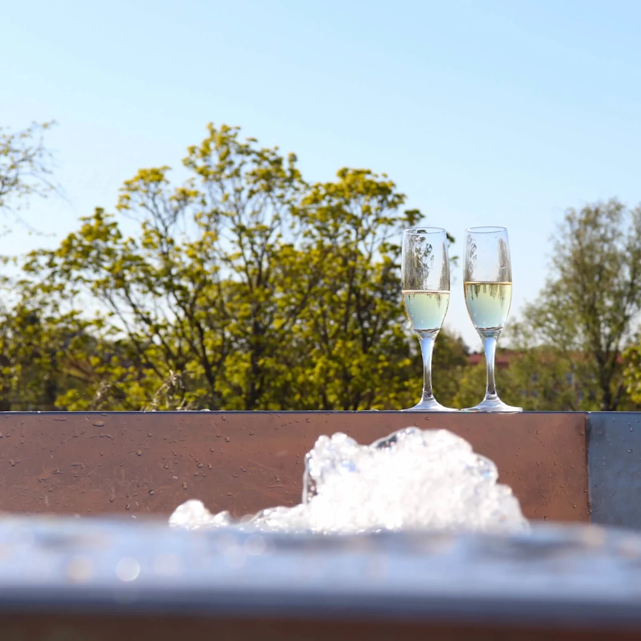 Två champagneglas, vilande på en våt yta med bubbligt skum, i en utomhusmiljö med gröna träd i bakgrunden.