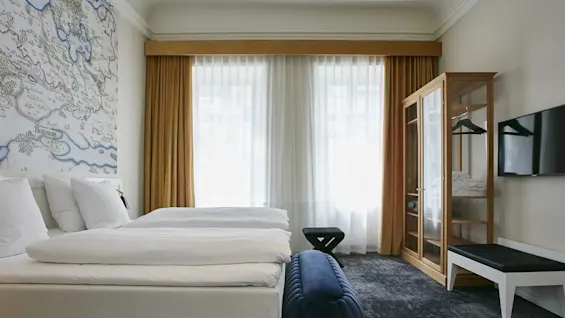 Et lyst og indbydende hotelværelse med to komfortable senge, et dekorativt vægmaleri og store vinduer.