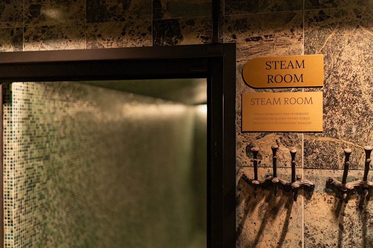 Sisäänkäynti höyryhuoneeseen, jossa kyltit lukevat 'Höyryhuone' ja kuvaavat sen etuja. Naulakot näkyvät kaakeloidulla seinällä.