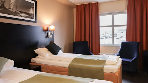 Two neatly made single beds with green runners are positioned beside a window. Two dark chairs and red curtains frame the white walls illuminated by a lamp, under a black-and-white photo.