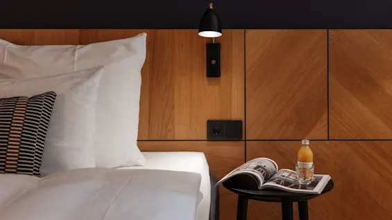 Et koselig hotellrom med en seng, en sengegavl i tre, en lampe og et sidebord med et magasin og juice.