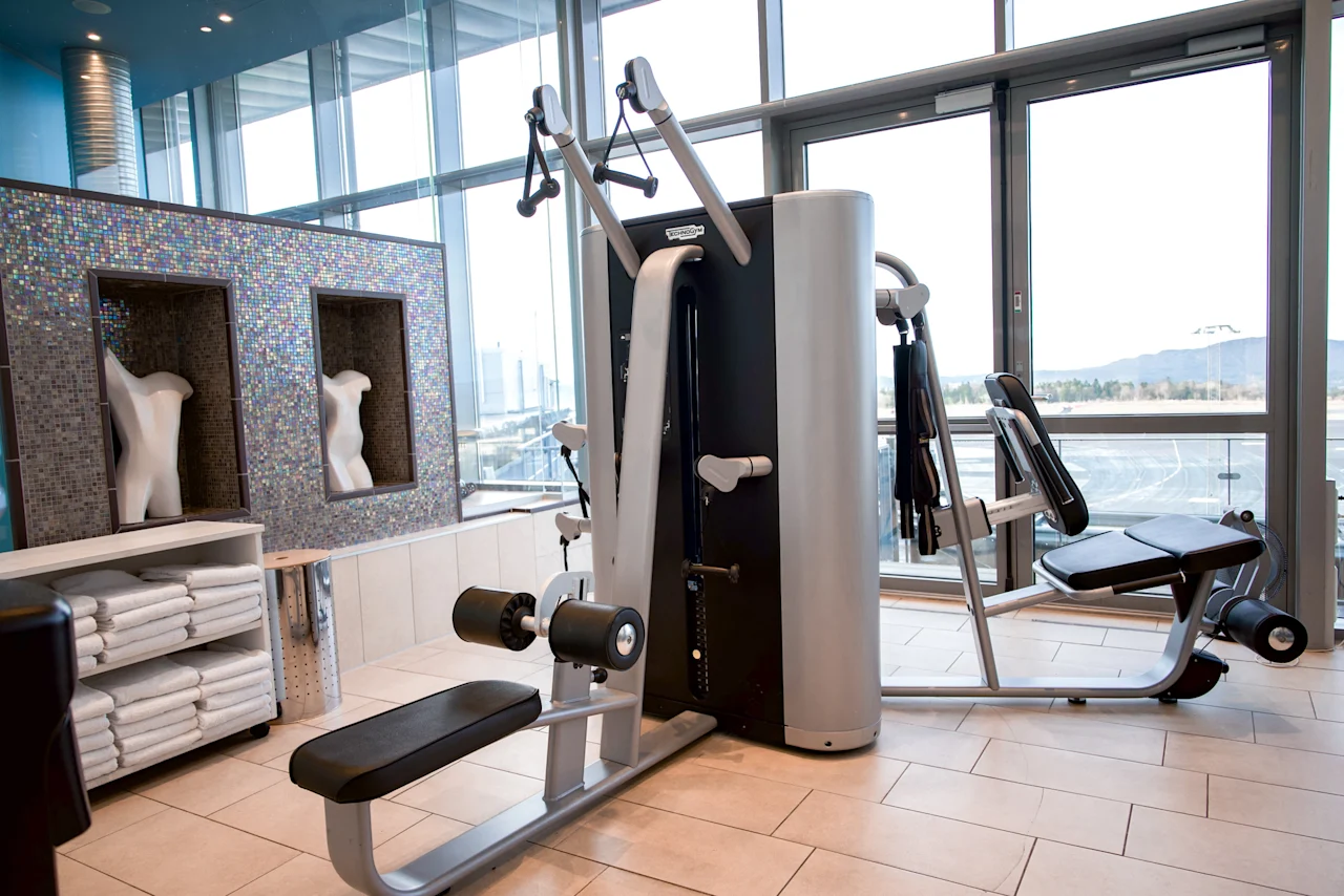En multigym-maskin med trinser og sæder står ubrugt i et moderne fitnesscenter. Den er omgivet af pænt stablede håndklæder, mosaikvægsudsmykning og store vinduer.