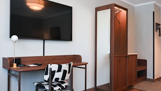 Et moderne hotellrom med skrivebord, stol, veggmontert TV og en garderobe i tre med helfigurspeil.
