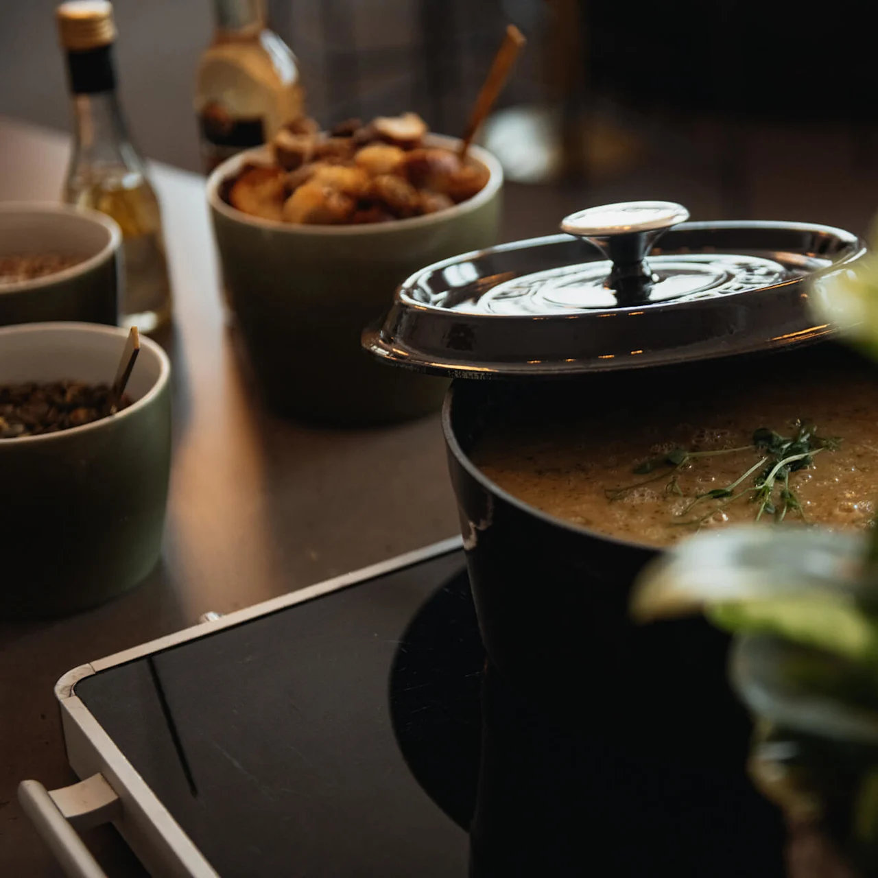 A pot of soup simmers on a stove, partially lidded, garnished with herbs. Nearby, bowls of seeds and roasted vegetables sit and a plant decorates the scene.